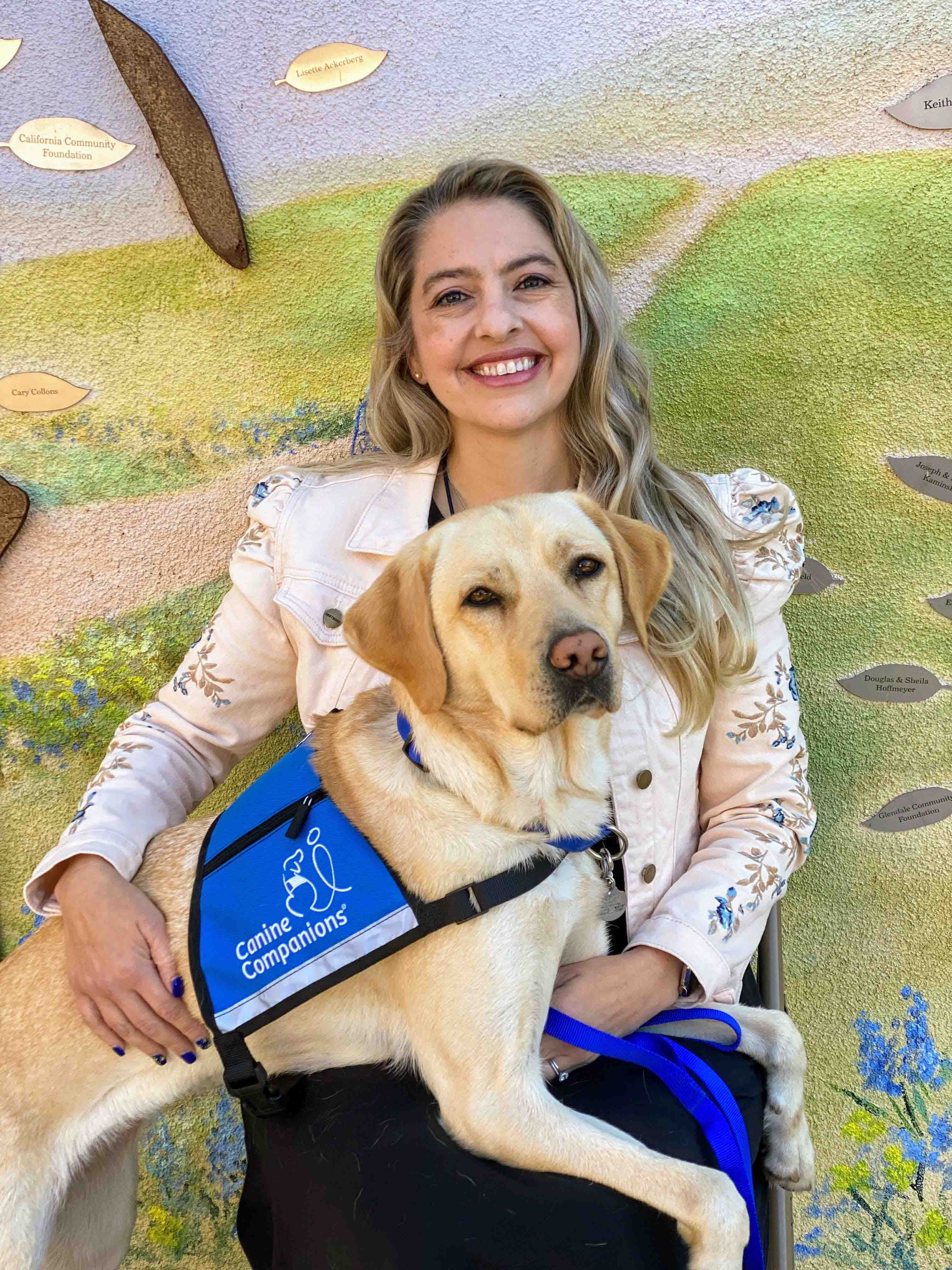 A white woman sits with a yellow lab weating a companion vest in her lap