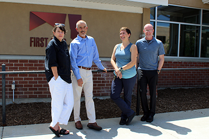 Four white adults stand in front of a building on a sunny summertime day; some of the group are looking at the camera, some are looking at each other